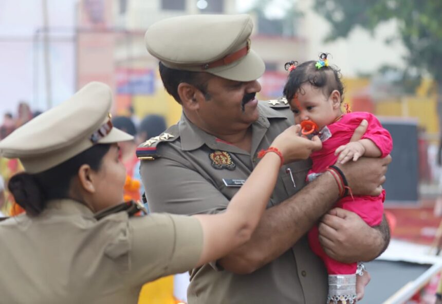 खनऊ के छठ घाट पर ड्यूटी के दौरान एक पुलिसकर्मी गोद में छोटे बच्चे को संभालते हुए, जबकि उसकी माँ पूजा कर रही है — भीड़ के बीच ममता और इंसानियत का सुंदर दृश्य।