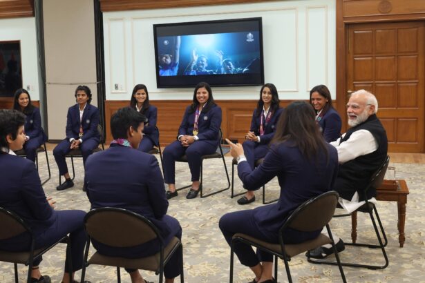 प्रधानमंत्री नरेंद्र मोदी भारतीय महिला क्रिकेट टीम के साथ वर्ल्ड कप ट्रॉफी पकड़े हुए, खिलाड़ी साइन की हुई जर्सी भेंट करते हुए।