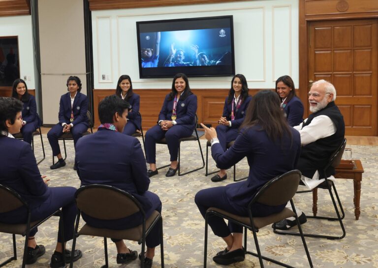 प्रधानमंत्री नरेंद्र मोदी भारतीय महिला क्रिकेट टीम के साथ वर्ल्ड कप ट्रॉफी पकड़े हुए, खिलाड़ी साइन की हुई जर्सी भेंट करते हुए।